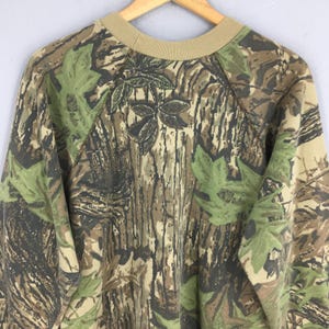 Op de afbeelding: Een sweatshirt met camouflagepatroon met een beige ronde hals en blad- en boomstamontwerpen. De top met lange mouwen heeft een bruin, groen en beige kleurenschema en hangt aan een houten hanger.