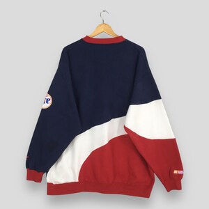 May include: A vintage color-block sweatshirt in navy, white, and red. The long-sleeved top features a crew neck with a red trim and a logo on the left sleeve. The back of the sweatshirt is visible, hanging from a wooden hanger.
