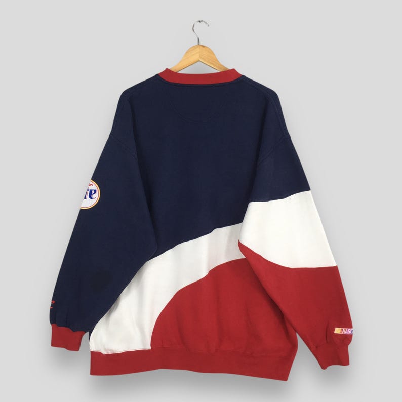 May include: A vintage color-block sweatshirt in navy, white, and red. The long-sleeved top features a crew neck with a red trim and a logo on the left sleeve. The back of the sweatshirt is visible, hanging from a wooden hanger.
