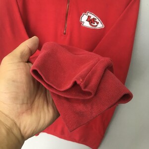 Op de afbeelding: Een felrood sweatshirt met lange mouwen, een kwartritssluiting bij de hals en een witte, pijlpuntvormige patch op de borst met de rode letters KC erin gedrukt. Het kledingstuk heeft geribbelde boorden en een zachte, effen gekleurde afwerking.