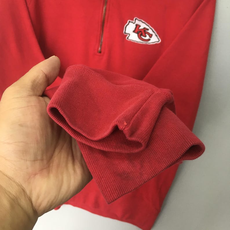 Op de afbeelding: Een felrood sweatshirt met lange mouwen, een kwartritssluiting bij de hals en een witte, pijlpuntvormige patch op de borst met de rode letters KC erin gedrukt. Het kledingstuk heeft geribbelde boorden en een zachte, effen gekleurde afwerking.