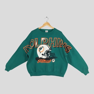 Puede incluir: Sudadera verde azulado con la palabra "DOLPHINS" en letras naranjas. Un casco de fútbol americano blanco con el logotipo del equipo está centrado debajo de la palabra. La palabra "Miami" está impresa en naranja debajo del casco.