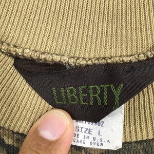 Op de afbeelding: Close-up van een bruin en groen camouflage-patroon shirt tag. De tag is zwart met het woord "LIBERTY" in groene letters. Een wit label hieronder geeft maat L en "MADE IN U.S.A."