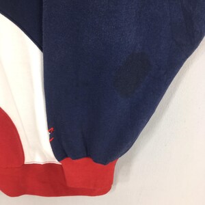 May include: Close-up of a section of a sweatshirt. The design features a color block pattern with navy blue, white, and red sections. The cuff is red. There is a dark stain on the navy blue fabric.