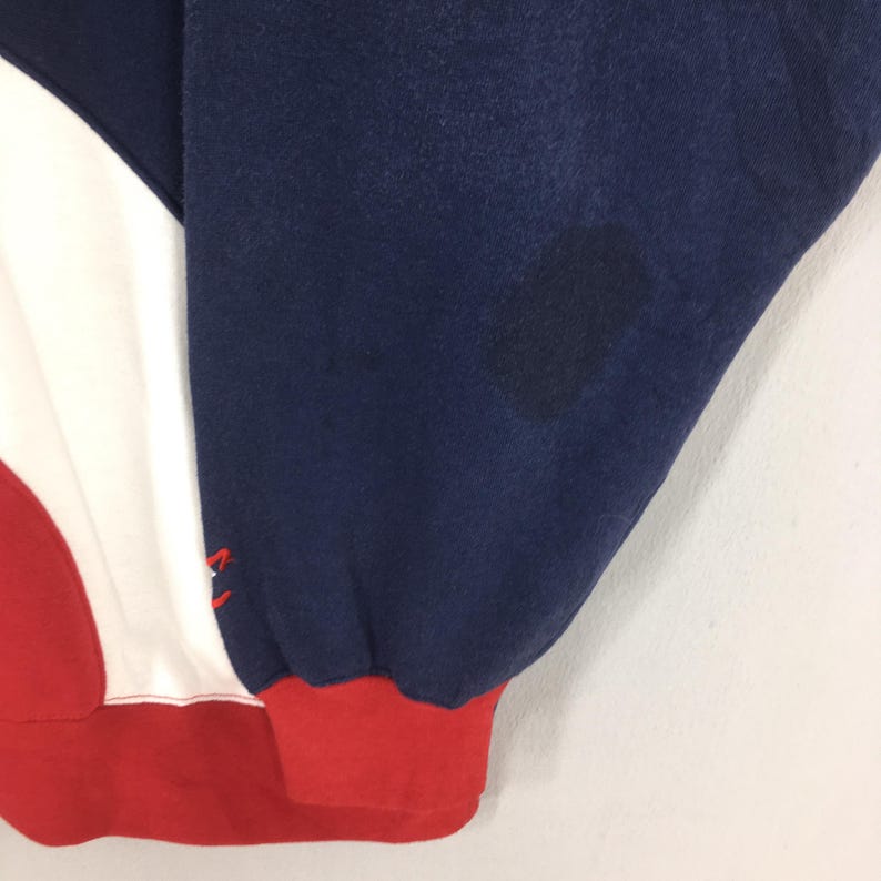 May include: Close-up of a section of a sweatshirt. The design features a color block pattern with navy blue, white, and red sections. The cuff is red. There is a dark stain on the navy blue fabric.
