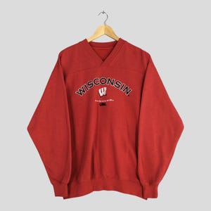 May include: A red v-neck sweatshirt with the word "WISCONSIN" arched across the chest in black lettering. Below the word "WISCONSIN" is a white "W" logo and the word "BADGERS" in black. The sweatshirt is hanging on a wooden hanger.