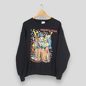 May include: Black sweatshirt with a colorful graphic of a skier holding skis and poles, with the word "PENNSYLVANIA" in red. The skier is wearing a hat, goggles, and a jersey with the number 92. White snowflake shapes are scattered around the graphic.