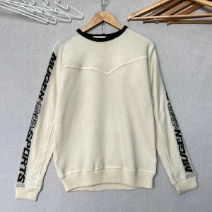 May include: A cream-coloured long-sleeve sweater featuring a black ribbed crew neckline and a V-shaped seam across the chest. The sleeves display the black text MUGEN SPORTS printed vertically along the outer arms, finished with ribbed cuffs.