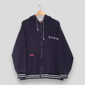 May include: A dark navy blue zip-up hoodie featuring a hood with drawstrings, long sleeves, and ribbed cuffs with two grey stripes. The chest displays small white graphic icons, and a small red rectangular tag is attached to the front pocket.