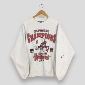 Op de afbeelding: Een lichtgrijs sweatshirt met een rood en wit grafisch ontwerp van een footballhelm en de woorden "National Champions". Het sweatshirt heeft ook het woord "Huskers" en de jaren 1994 en 1995.
