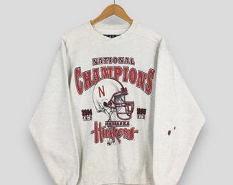 Peut inclure: Un sweat-shirt gris clair avec un graphisme rouge et blanc représentant un casque de football et les mots "National Champions". Le sweat-shirt comporte également le mot "Huskers" et les années 1994 et 1995.