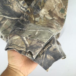 May include: Close-up of a camouflage-patterned sweatshirt sleeve. The fabric features a blend of brown, green, and tan hues, creating a woodland camouflage design. The cuff is visible, showing the fabric's texture and stitching.