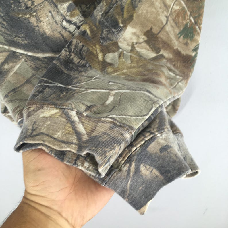 May include: Close-up of a camouflage-patterned sweatshirt sleeve. The fabric features a blend of brown, green, and tan hues, creating a woodland camouflage design. The cuff is visible, showing the fabric's texture and stitching.