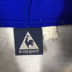 May include: Close-up of a blue and white athletic garment. A navy blue label with the text "le coq sportif" and a rooster logo is visible. The garment has a textured blue collar.