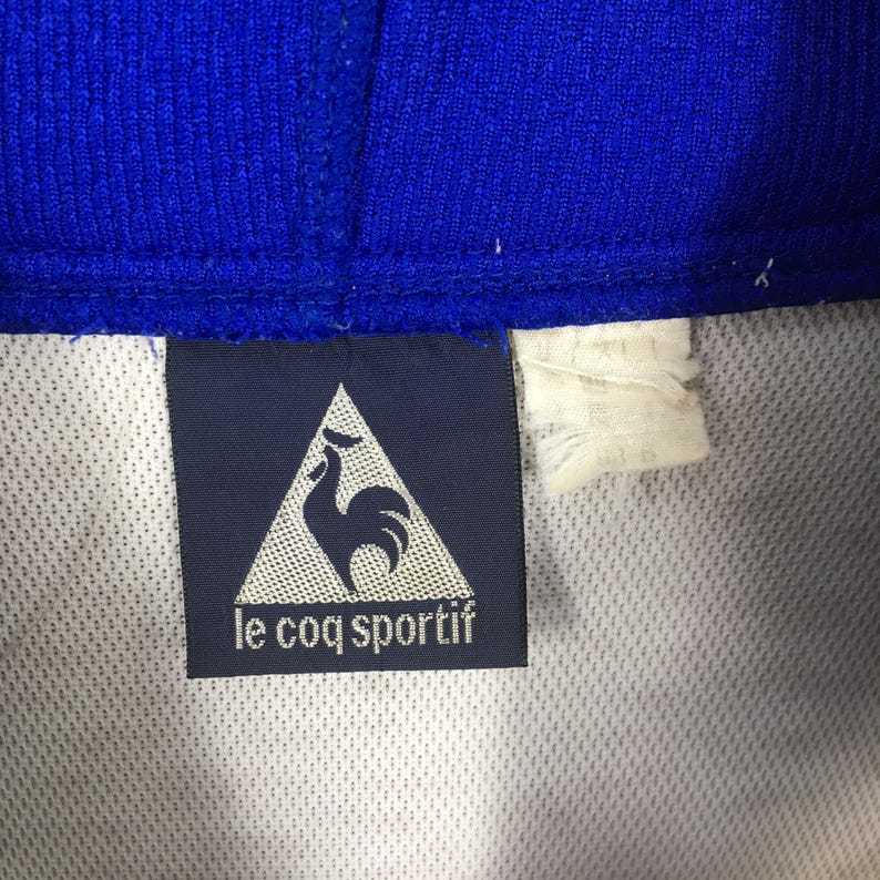 May include: Close-up of a blue and white athletic garment. A navy blue label with the text "le coq sportif" and a rooster logo is visible. The garment has a textured blue collar.