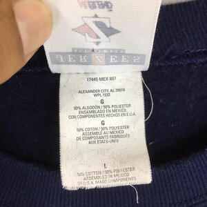 May include: Close-up of a navy blue garment tag with text in English and Spanish. The tag includes the text "ALEXANDER CITY, AL 35010" and "ASSEMBLED IN MEXICO". The tag also has a logo with a red, white, and blue design.
