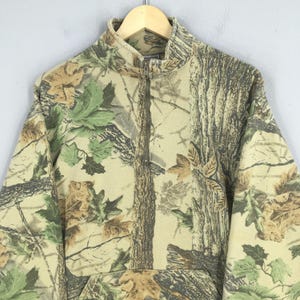 Puede incluir: Una sudadera con cremallera de cuarto de camuflaje. El dise&ntilde;o presenta una mezcla de tonos marrones, verdes y beiges, imitando un entorno forestal. La sudadera tiene un cuello alto y un bolsillo delantero.