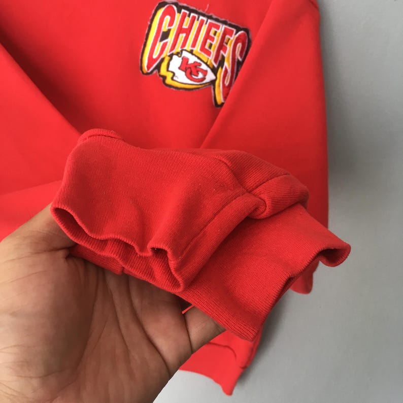 Peut inclure: Un sweat-shirt rouge vif &agrave; manches longues avec un poignet c&ocirc;tel&eacute; et un &eacute;cusson graphique sur la poitrine. L'&eacute;cusson affiche le mot CHIEFS en lettres jaunes et blanches audacieuses au-dessus d'un embl&egrave;me de logo d'&eacute;quipe en forme de pointe de fl&egrave;che.