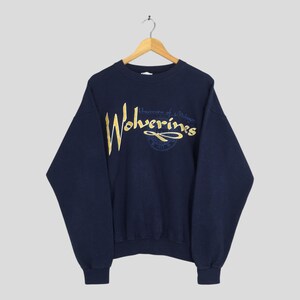 May include: Navy blue sweatshirt with gold embroidered text that reads "Wolverines" and "University of Michigan". The sweatshirt is hanging on a wooden hanger against a light gray background.