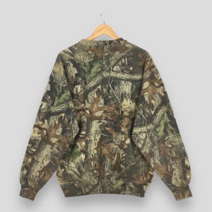 K&ouml;nnte beinhalten: Ein Sweatshirt mit Camouflage-Muster, Rundhalsausschnitt und langen &Auml;rmeln. Das Design zeigt eine Mischung aus Braun-, Gr&uuml;n- und Beiget&ouml;nen, die eine Waldumgebung nachahmen. Das Sweatshirt h&auml;ngt an einem Holzb&uuml;gel.