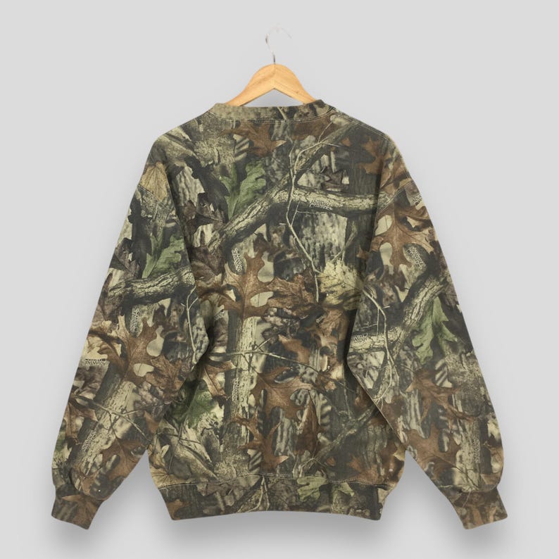 K&ouml;nnte beinhalten: Ein Sweatshirt mit Camouflage-Muster, Rundhalsausschnitt und langen &Auml;rmeln. Das Design zeigt eine Mischung aus Braun-, Gr&uuml;n- und Beiget&ouml;nen, die eine Waldumgebung nachahmen. Das Sweatshirt h&auml;ngt an einem Holzb&uuml;gel.
