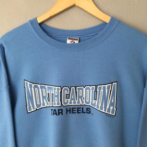 May include: Light blue sweatshirt with the words "NORTH CAROLINA" in white and blue block letters. Below the text is the phrase "TAR HEELS" in white letters. The sweatshirt has a crew neck and long sleeves.