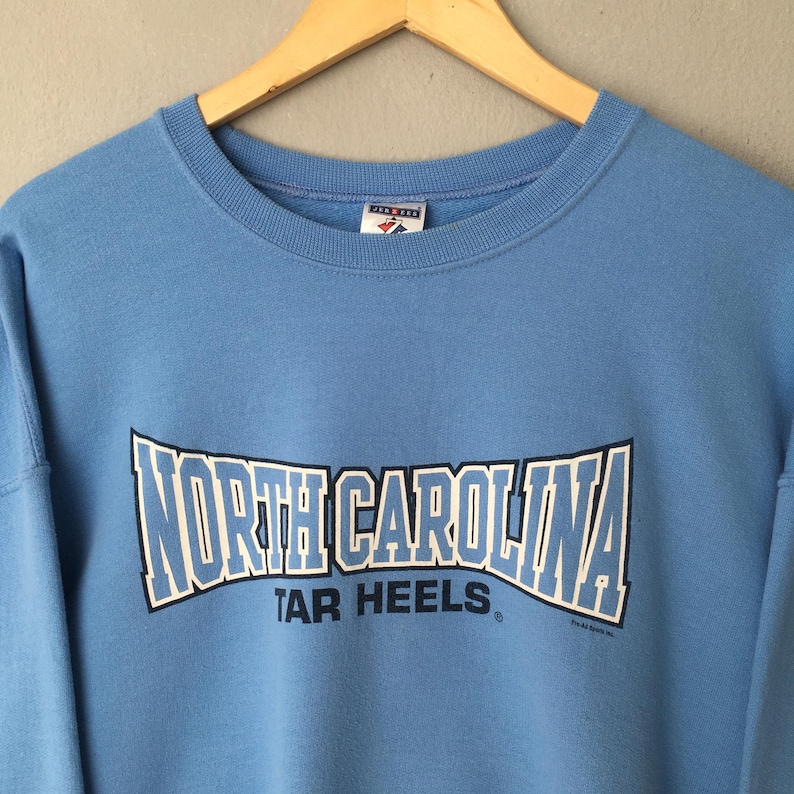 May include: Light blue sweatshirt with the words "NORTH CAROLINA" in white and blue block letters. Below the text is the phrase "TAR HEELS" in white letters. The sweatshirt has a crew neck and long sleeves.