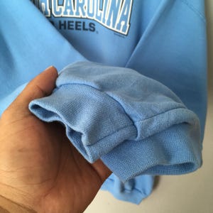 May include: A light blue sweatshirt with the text "NORTH CAROLINA" arched across the top and "HEELS" below. The cuff of the sleeve is visible, showing the ribbed knit fabric.