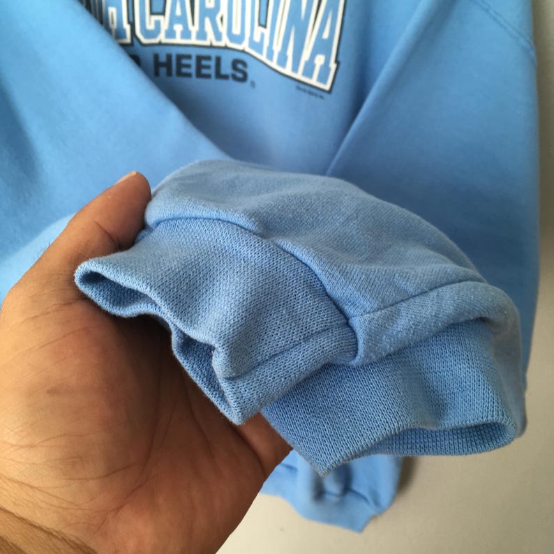 May include: A light blue sweatshirt with the text "NORTH CAROLINA" arched across the top and "HEELS" below. The cuff of the sleeve is visible, showing the ribbed knit fabric.