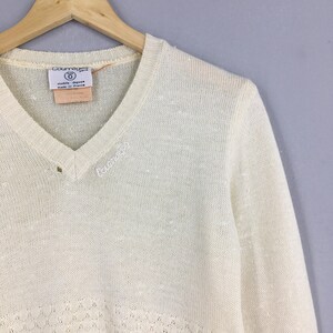 May include: Cream-colored Courr&egrave;ges Paris V-neck sweater. The sweater has a subtle knit pattern at the bottom and the Courr&egrave;ges logo embroidered on the chest. A tag with the Courr&egrave;ges logo and text is visible near the neckline. The sweater is hanging on a wooden hanger.