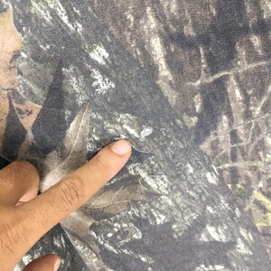 Puede incluir: Primer plano de una tela con estampado de camuflaje. La tela presenta una mezcla de tonos marrones, verdes y grises, creando un dise&ntilde;o naturalista de bosque. Un dedo est&aacute; tocando la tela.