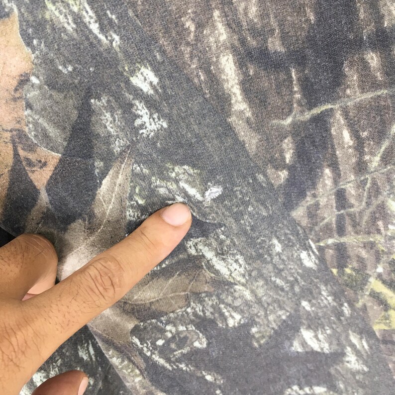 Puede incluir: Primer plano de una tela con estampado de camuflaje. La tela presenta una mezcla de tonos marrones, verdes y grises, creando un dise&ntilde;o naturalista de bosque. Un dedo est&aacute; tocando la tela.