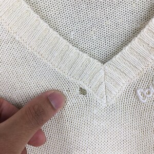 May include: Close-up of an off-white V-neck sweater. The sweater has a ribbed neckline and a small hole near the collar. The knit fabric is visible, and the texture is slightly uneven.