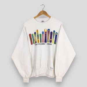 Könnte beinhalten: Weißes Sweatshirt mit einer Grafik von bunten Snowboards. Die Snowboards sind in einer Reihe angeordnet, mit verschiedenen Designs und Farben. Der Text "PRAY FOR SNOW • HAWAII" ist darunter aufgedruckt. Das Sweatshirt hat lange Ärmel und einen Rundhalsausschnitt.
