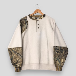 May include: A light beige sweatshirt with long sleeves and a crew neck, featuring a three-button placket. The sleeves and shoulder areas have a camouflage pattern in shades of brown, green, and tan. The sweatshirt is hanging on a wooden hanger.