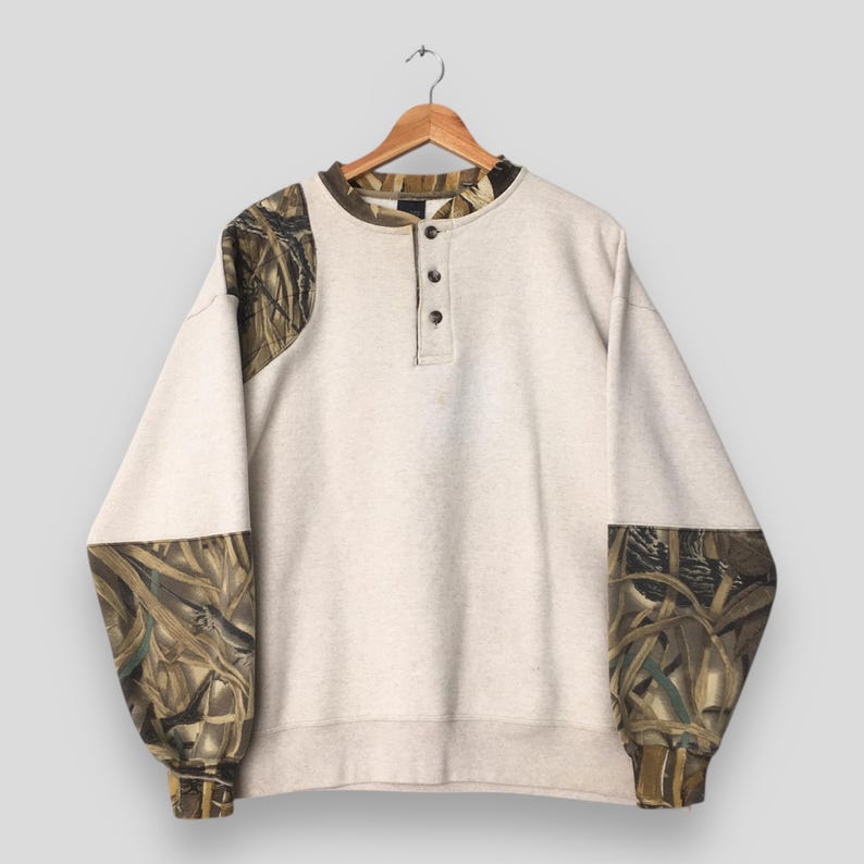 May include: A light beige sweatshirt with long sleeves and a crew neck, featuring a three-button placket. The sleeves and shoulder areas have a camouflage pattern in shades of brown, green, and tan. The sweatshirt is hanging on a wooden hanger.