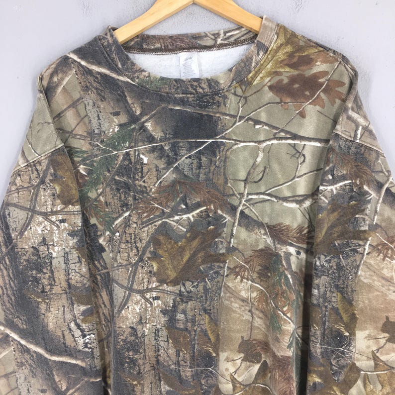 May include: A long-sleeved camouflage sweatshirt with a crew neck. The pattern features shades of brown, green, and tan, mimicking a forest environment. The sweatshirt is hanging on a wooden hanger.