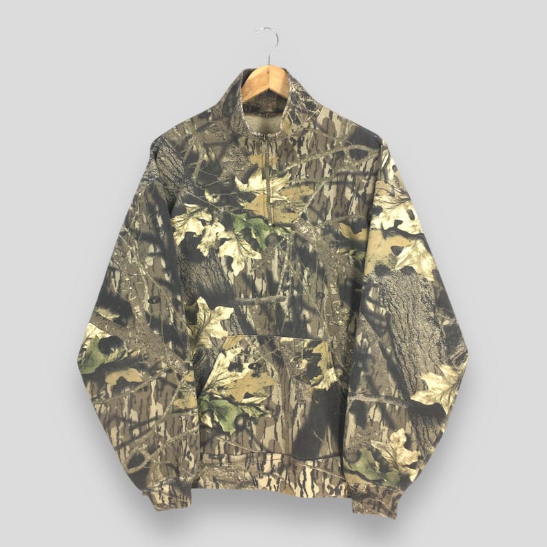 Pode incluir: Um moletom pullover com estampa camuflada, fecho de z&iacute;per de um quarto e bolso canguru. O design incorpora tons de marrom, verde e bege, imitando um ambiente florestal. O moletom est&aacute; pendurado em um cabide de madeira.