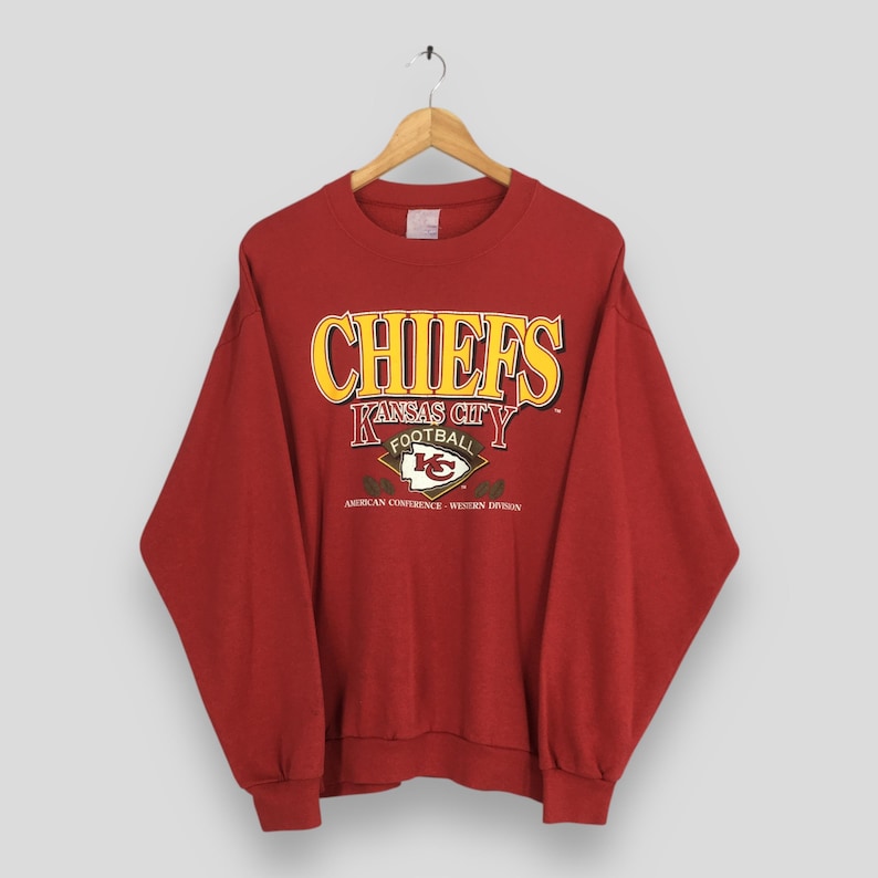 May include: A red crewneck sweatshirt featuring the text "CHIEFS" in yellow and white, with "KANSAS CITY FOOTBALL" below. The sweatshirt also displays the team logo and "AMERICAN CONFERENCE - WESTERN DIVISION".