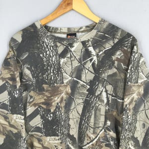 May include: A camouflage-patterned sweatshirt in shades of brown, gray, and green, hanging on a wooden hanger. The crew neck sweatshirt features a forest-like design, with tree and leaf patterns. The garment is made of a soft material.