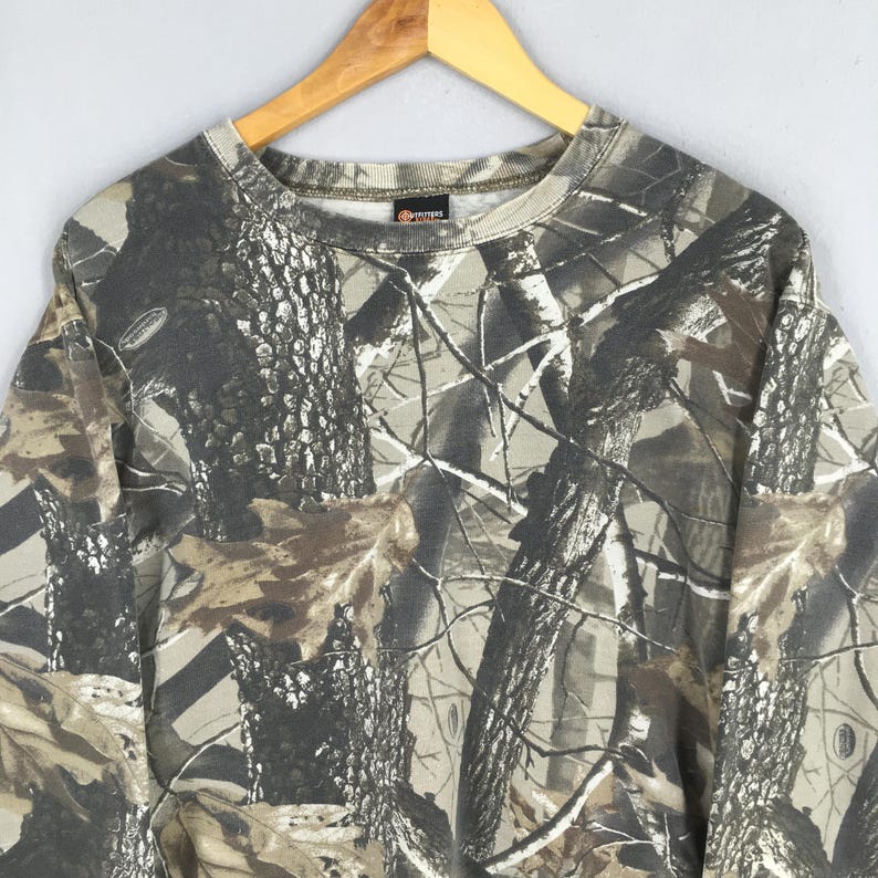 May include: A camouflage-patterned sweatshirt in shades of brown, gray, and green, hanging on a wooden hanger. The crew neck sweatshirt features a forest-like design, with tree and leaf patterns. The garment is made of a soft material.