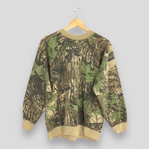 Op de afbeelding: Een sweatshirt met camouflagepatroon met een ronde hals en lange mouwen. Het ontwerp heeft een mix van bruine, groene en beige kleuren, die lijken op een boslandschap. De manchetten en de halslijn zijn effen beige van kleur. De sweatshirt hangt aan een houten hanger.