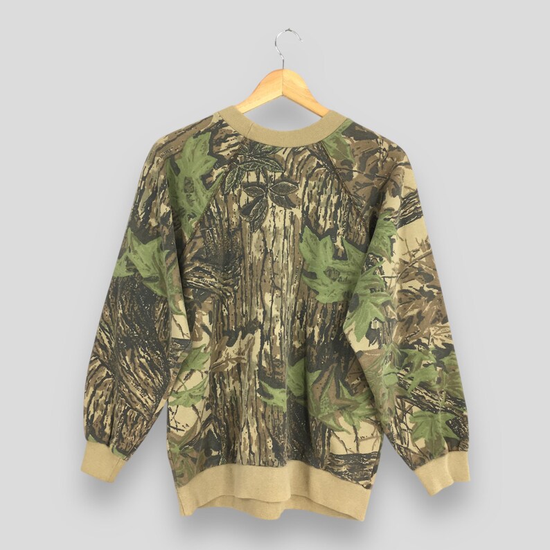 Op de afbeelding: Een sweatshirt met camouflagepatroon met een ronde hals en lange mouwen. Het ontwerp heeft een mix van bruine, groene en beige kleuren, die lijken op een boslandschap. De manchetten en de halslijn zijn effen beige van kleur. De sweatshirt hangt aan een houten hanger.