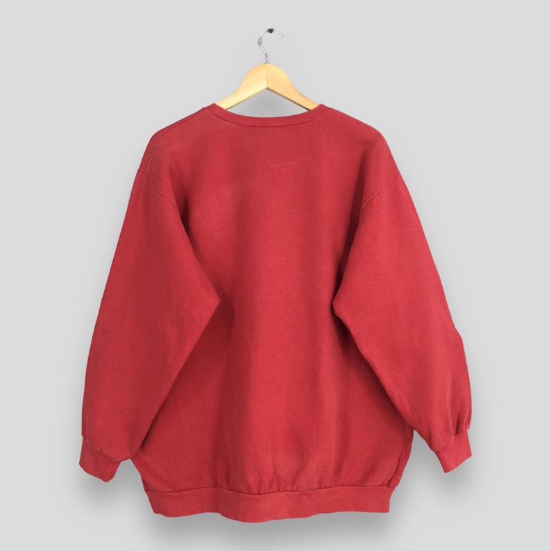 May include: A solid red sweatshirt hangs from a wooden hanger against a light gray background. The crew neck sweatshirt has long sleeves and a ribbed hem and cuffs. The fabric appears to be a soft, slightly faded material.