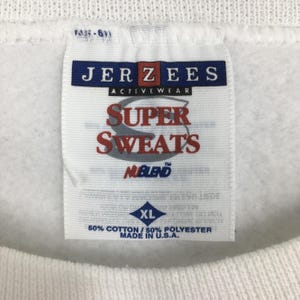 Może przedstawiać: Biała metka bluzy Jerzees Super Sweats NUBLEND z diamentowatą metką rozmiaru XL. Metka zawiera napis "50% bawełny / 50% poliester Wyprodukowano w USA."