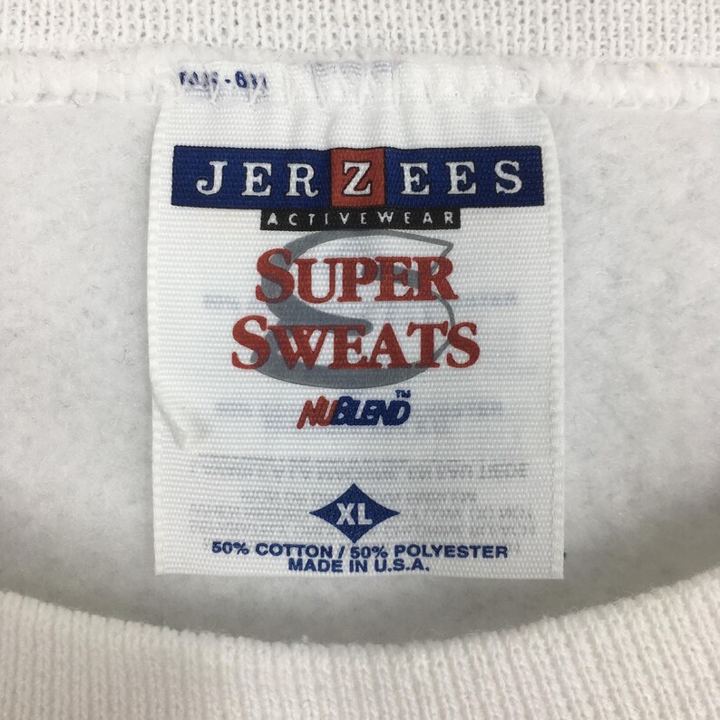 Może przedstawiać: Biała metka bluzy Jerzees Super Sweats NUBLEND z diamentowatą metką rozmiaru XL. Metka zawiera napis "50% bawełny / 50% poliester Wyprodukowano w USA."