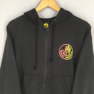 May include: Black zip-up hoodie with a hood and front pockets. The hoodie features a circular logo on the chest with a hand design and the words "Body Glove" in yellow and orange. The hoodie is hanging on a wooden hanger.