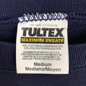 May include: Close-up of a navy blue sweatshirt tag. The tag reads "TULTEX MAXIMUM SWEATS" and lists the fabric composition as 50% cotton and 50% polyester. It also indicates the garment was assembled in Mexico. The size is Medium.