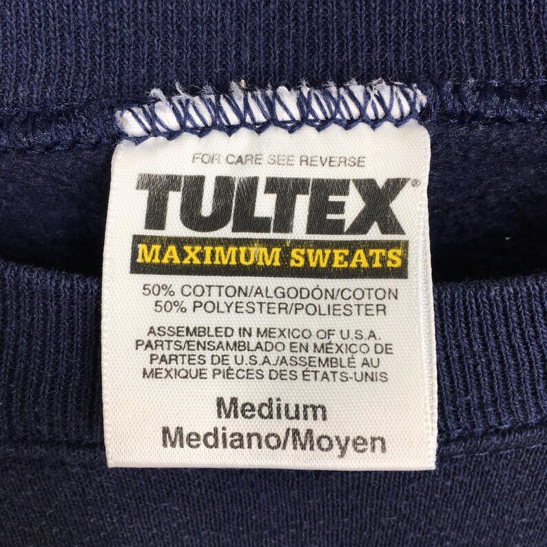 May include: Close-up of a navy blue sweatshirt tag. The tag reads "TULTEX MAXIMUM SWEATS" and lists the fabric composition as 50% cotton and 50% polyester. It also indicates the garment was assembled in Mexico. The size is Medium.