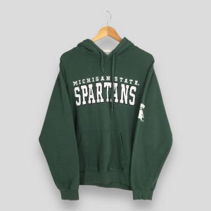 Puede incluir: Una sudadera con capucha verde oscuro con las palabras "MICHIGAN STATE" encima de "SPARTANS" en letras blancas. La sudadera tiene un bolsillo delantero y una capucha con cordón. Un pequeño gráfico blanco está en la manga derecha.
